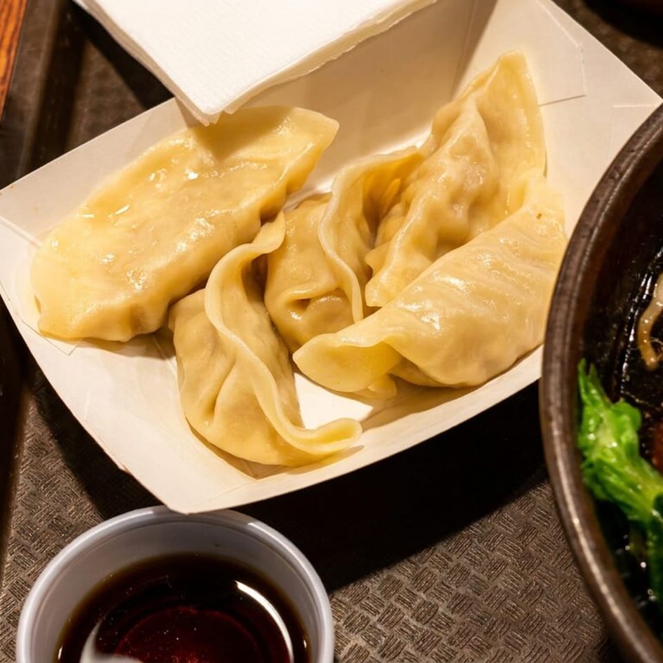 Steamed dumplings, ramen with tomato and greens, soy sauce, and water on a tray.